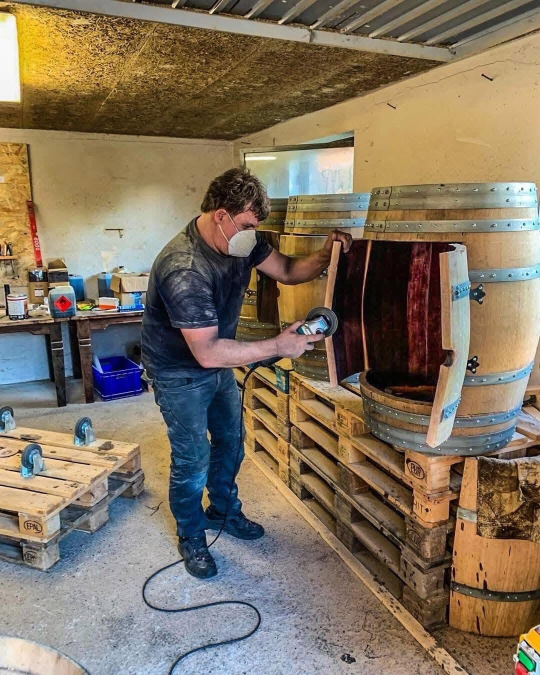 Man wearing a dust mask sanding a cut-open wine barrel in the workshop, more barrels stacked on pallets beside him