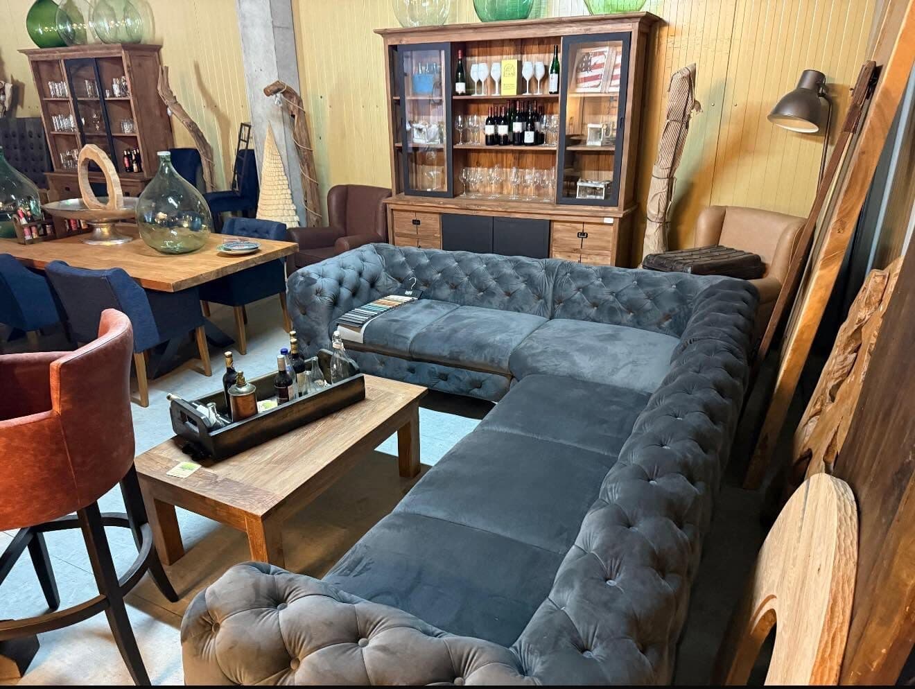 Blue-grey buttoned Chesterfield corner sofa in the showroom, a glass-front buffet cabinet behind it